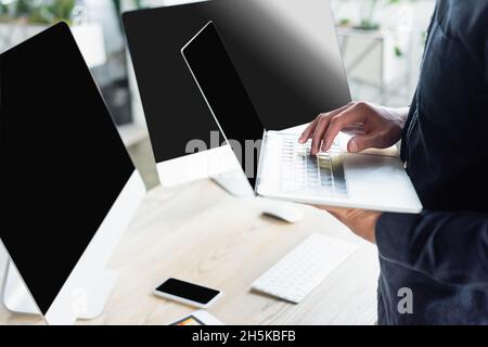 Cropped view of programmer using laptop near computers with blank screen in office Stock Photo