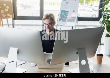 Happy developer in eyeglasses looking at computer near laptop and smartphone in office Stock Photo