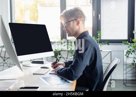 Side view of developer writing on notebook near computers with blank screen and smartphone in office Stock Photo