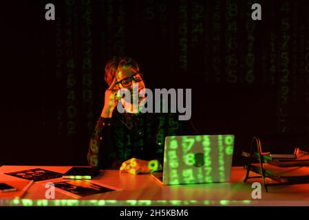 Smiling programmer in eyeglasses looking at laptop near reflection of computer code on black background Stock Photo