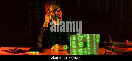 Smiling programmer in eyeglasses looking at laptop with reflection of computer code near papers on black background, banner Stock Photo