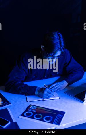 Programmer in eyeglasses writing on notebook near papers and devices on table isolated on black Stock Photo