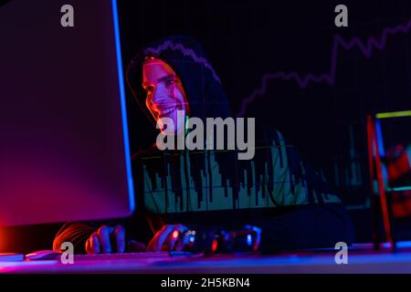 Smiling hacker using computer near eyeglasses and reflection of charts on black background Stock Photo