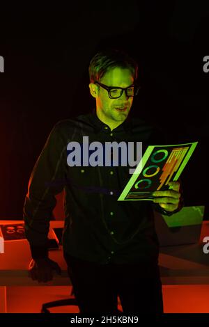 Smiling programmer holding document with charts near laptop with reflection on black background Stock Photo