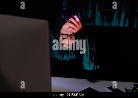 Sad hacker with reflection of charts sitting near devices on black background Stock Photo