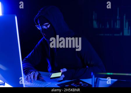 Hacker in balaclava using computer and smartphone on black background with charts reflection Stock Photo