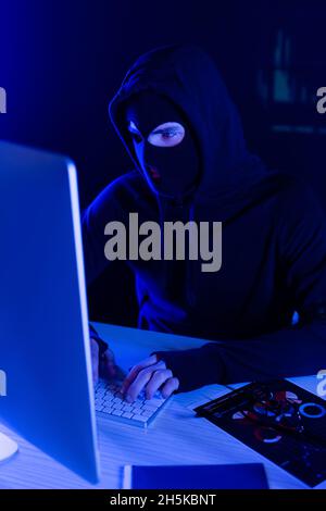 Hacker in balaclava using computer near papers on table on black background Stock Photo