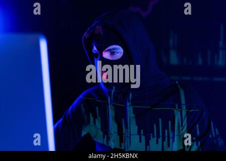 Hacker in balaclava with charts reflection looking at blurred computer monitor on black background Stock Photo