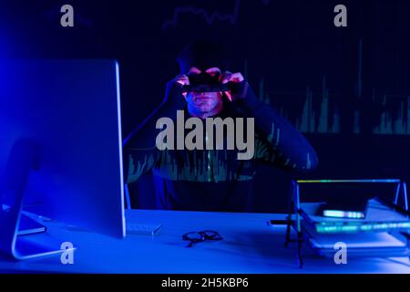 Hacker wearing balaclava near smartphone and computer on black background with charts reflection Stock Photo