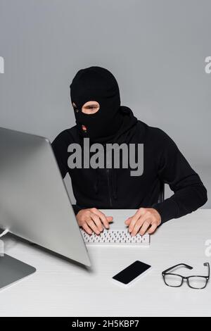 Hacker in balaclava using computer near smartphone and eyeglasses isolated on grey Stock Photo
