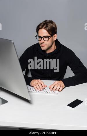 Programmer in eyeglasses using computer near smartphone isolated on grey Stock Photo