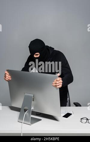 Hacker in balaclava taking computer monitor near smartphone isolated on grey Stock Photo