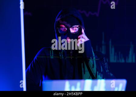 Pensive hacker in balaclava looking at laptop on black background with charts reflection Stock Photo