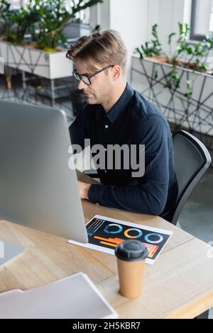 Programmer in eyeglasses using computer near document and coffee in office Stock Photo