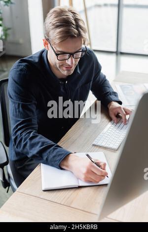 Programmer in eyeglasses writing on notebook and using computer in office Stock Photo