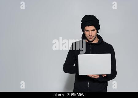 Hacker in balaclava using laptop on grey background Stock Photo