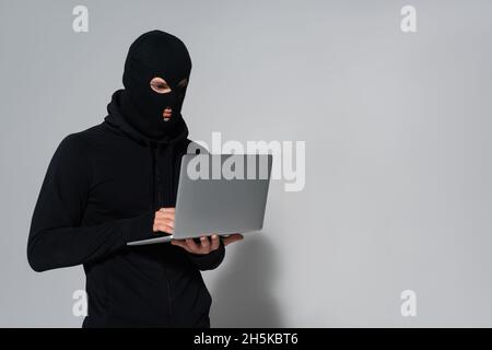 Hacker in balaclava using laptop on grey background Stock Photo