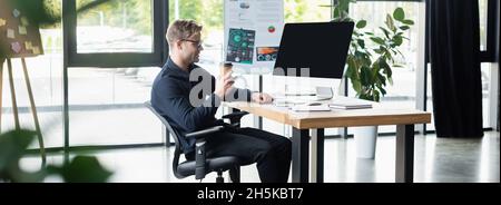 Side view of smiling programmer holding takeaway drink near computer with blank screen in office, banner Stock Photo