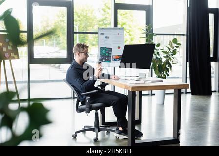 Side view of smiling developer holding coffee to go near computer monitor in office Stock Photo