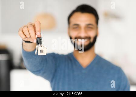 Selective focus on the keys with keychain in form of little house in male hand. Proud Indian man holds keys from new property, happy buyer of an own estate. Happy Hispanic guy rent new apartment Stock Photo