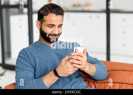 Focused bearded Indian man lying on the sofa at home and holding smartphone, Hispanic guy reading news online, spend leisure time in social networks, dark-haired arab male texting and chatting online Stock Photo