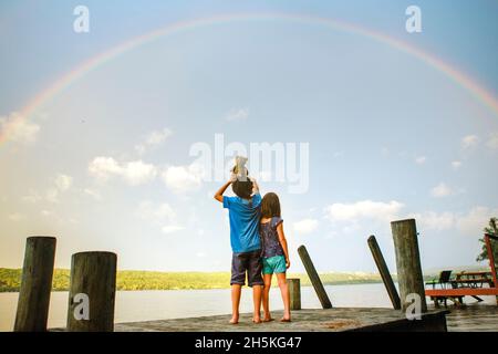 two children stand on deck under rainbow by sunlit lake with toy bear Stock Photo