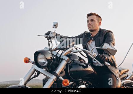 Stylish young attractive guy is sitting on a motorcycle. A handsome young man in a black leather jacket is driving a black bike. Sunny evening. Stock Photo