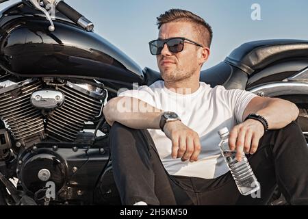 Young attractive guy in sunglasses is holding a bottle of clean water. A nice young man in a white T-shirt sits and rests on the background of a motor Stock Photo