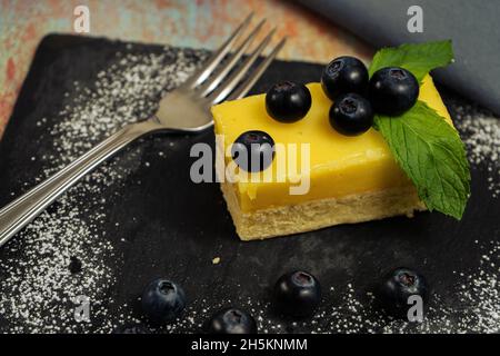 One Portion of homemade lemon curd cake in the shape of an ingot or brick decorated with some mint leaves and blueberries on a black slab. High view. Stock Photo