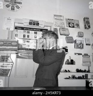 1960, historial, a man holding a camera in a electrcial shop, England, UK, looking through the viewfinder, perhaps a customer testing it with a new battery. On the wall, the traditional peg board, used to display smaller electric items; irons, lights, clocks, torches, plugs. Famous names of the era can be seen; Ronson, Ever Ready.  A promotional poster for the Dynatron range of high quality Radiogram and wirelss products is displayed. A British radio manufacturer, Dynatron was established in 1927 by the Hacker brothers, Ron and Arthur, above the family grocery shop on Maidenhead High Street. Stock Photo