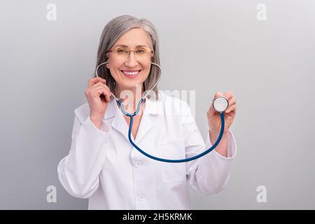 Photo of pretty charming mature lady doctor wear white coat spectacles smiling listening your heartbeat isolated grey color background Stock Photo