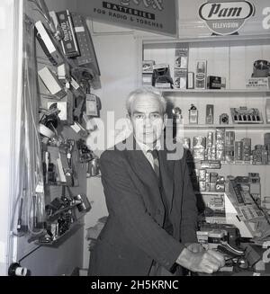 1960, a male shopkeeper at the counter of his electrical goods store, England, UK. Behind his right shoulder on the wall a traditional peg board, used to display smaller items; irons, lights, clocks, torches, plugs. On wooden shelves, a promotional card for Pam, a brand name of British manufacturer Pamphonic. Founded by Paul Taylor and Edgar Lavington in 1932 in the garage of his mother's house in Prince Albert St, London, Pamphonoc Reproducers Ltd made high quality sound systems. 1n 1947 Pye brought control. Stock Photo