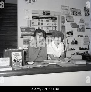 1960s, historical, female shopkeeper and lady assistant standing at the counter in an electrical goods store, England, UK. Behind them on the walls, the traditional peg board, used to display smaller items; irons, lights, clocks, torches, plugs. Famous names of the era can be seen, including Ronson, Every Ready, Exide, Pifer. A promotional poster for the Dynatron range of high quality Radiogram and wirelss products is displayed. A British radio manufacturer, Dynatron was established in 1927 by the Hacker brothers, Ron and Arthur, above the family grocery shop on Maidenhead High Street. Stock Photo