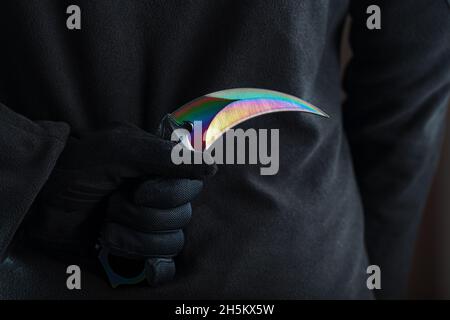 The man in black holds a curved kerambit knife behind his back. A criminal with a knife hidden behind his back Stock Photo