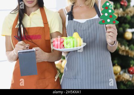Female waiters holding plate of fruit and writing down order near New Year tree closeup Stock Photo