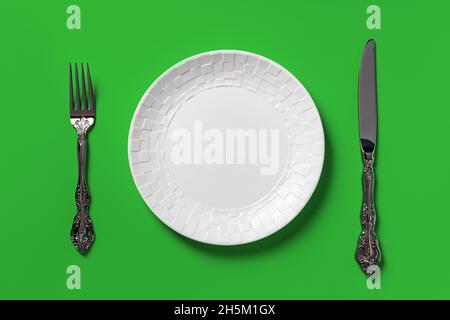 Empty white dinner plate, fork and table knife against green background. Blank mockup template for dieting, healthy eating and good eating habits. Stock Photo
