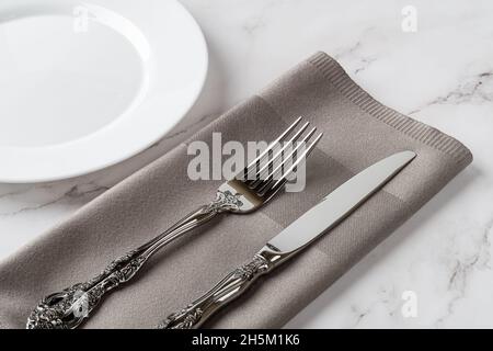 Elegant vintage fork and table knife on a gray dinner napkin and white porcelain plate over marble tabletop. Crockery and cutlery for set table. Empty Stock Photo