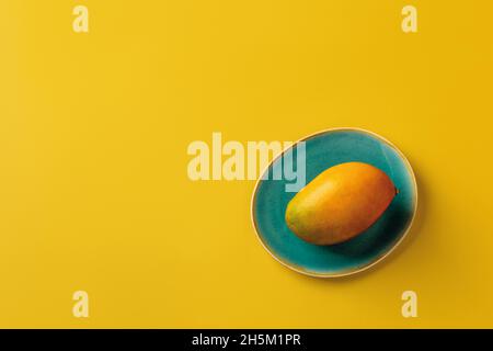 Whole ripe yellow Palmer mango on the turquoise plate over bright yellow background. Ready to eat sweet delicious summer fruit of Mangifera Indica. Stock Photo