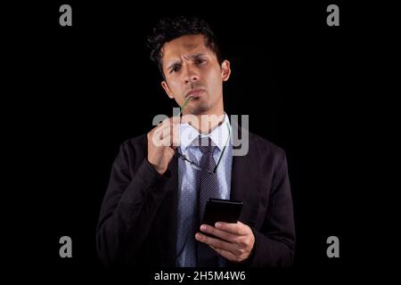 Businessman with cell phone in hand deep in thought while looking straight ahead. Young latin businessman with glasses in his hand while thinking of a Stock Photo