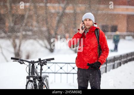 Caucasian man in red jacket calling with his smartphone, standing close to his bike at winter season Stock Photo