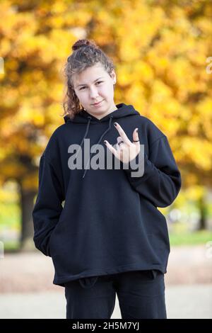 Portrait of young woman gesturing cool with hand, black hoody and trendy jeans, autumn season with yellow leaves Stock Photo