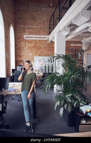 Full length shot of cheerful woman, architect or interior designer smiling away while talking on the phone, standing in modern green office Stock Photo
