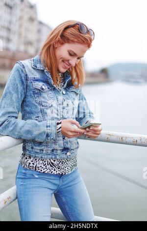 Young redhead cheerful Caucasian woman messaging on smartphone at riverbank Stock Photo