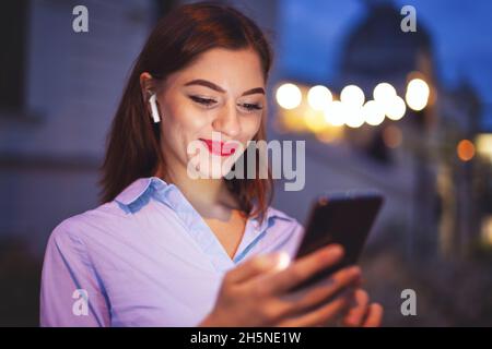 Young cheerful redhead businesswoman checking messages on smartphone at night in city Stock Photo