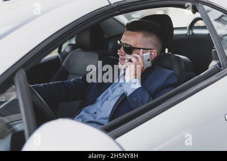 Portrait of handsome businessman speaking by phone sitting in seat of car and smiling happily. driver communicates on a smartphone while driving. busi Stock Photo