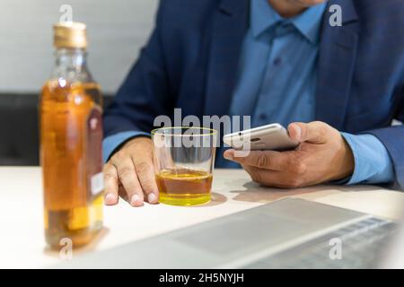 A businessman drinks alcohol and looks at information through his smartphone while sitting at a table next to a laptop in a bar. Drink alcohol in the Stock Photo