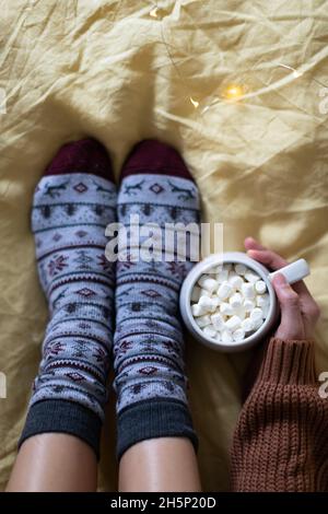 A large mug of cocoa in the hands of a girl in a warm sweater, sitting in warm socks on the bed. Top view. Stock Photo