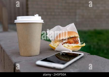 A paper cup with coffee or tea, a chicken sandwich and a smartphone are lying on a bench on the street. The concept of junk food on the lunch break Stock Photo