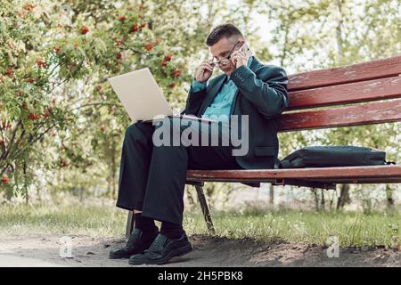 Handsome man working on his laptop. businessman sitting on the bench and watching on the laptop in city park outdoors. Internet connection allows peop Stock Photo