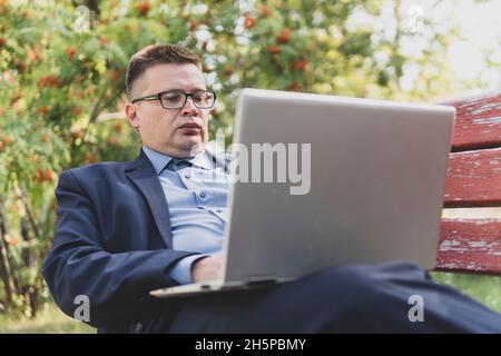 Satisfied businessman working on laptop with internet wireless. male programmer works against the background of a green park in nature. Stock Photo